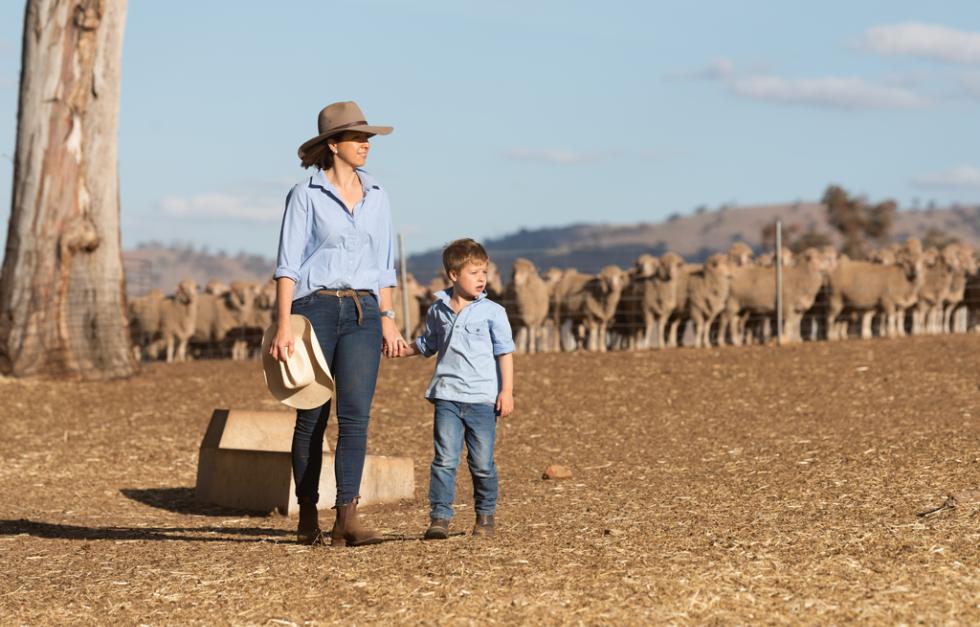 Woman and child standing on dry farm paddock with sheep in background