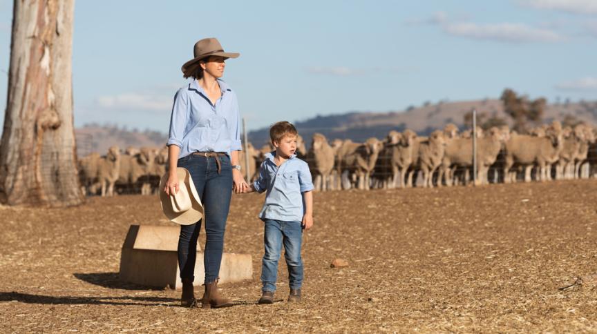 Woman and child standing on dry farm paddock with sheep in background