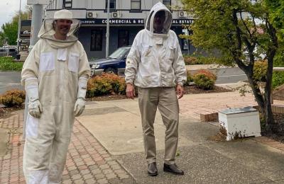 Two people wearing beekeeping suits standing beside bee box. 